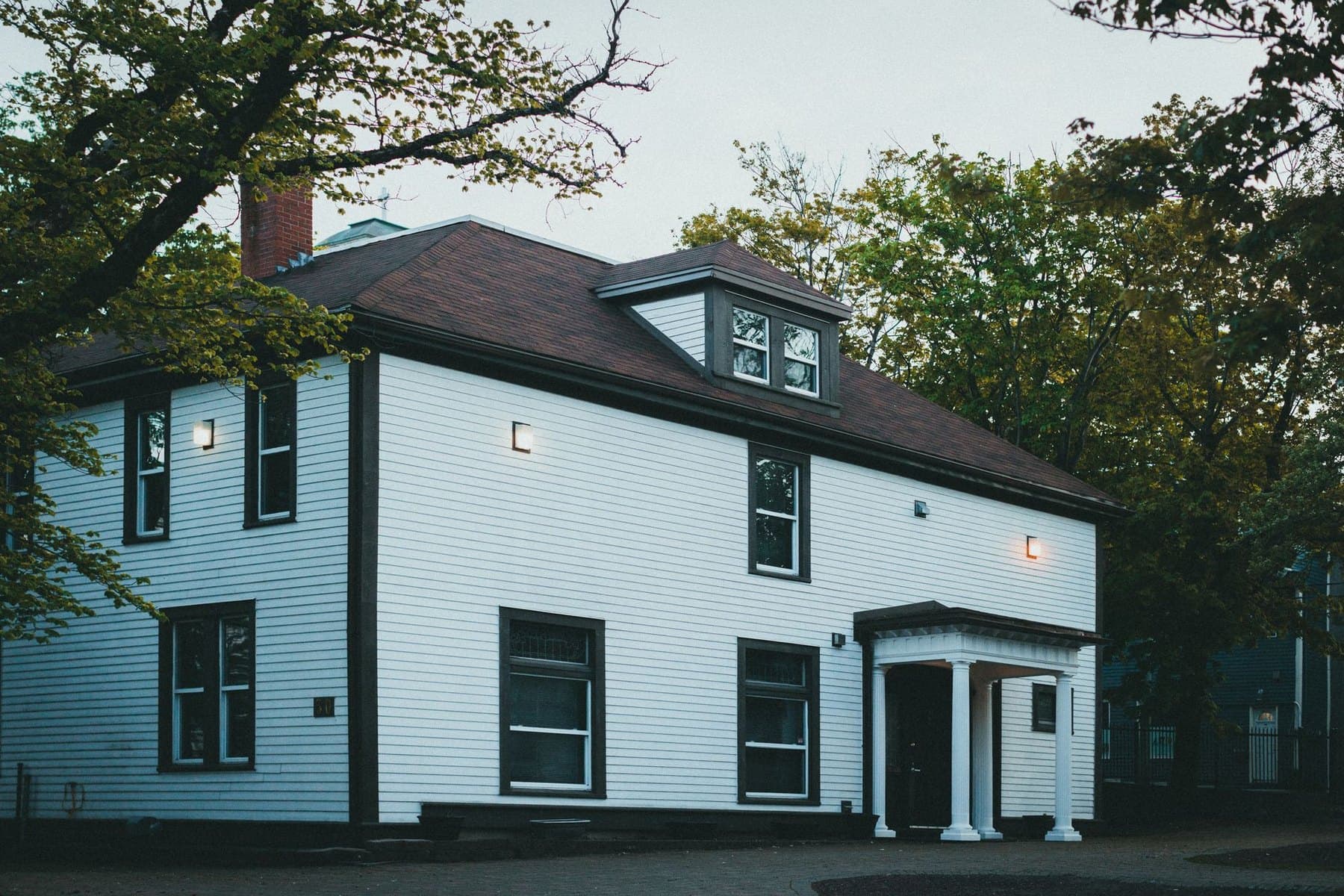 A white clapboard family home among mature trees, lit from inside at dusk