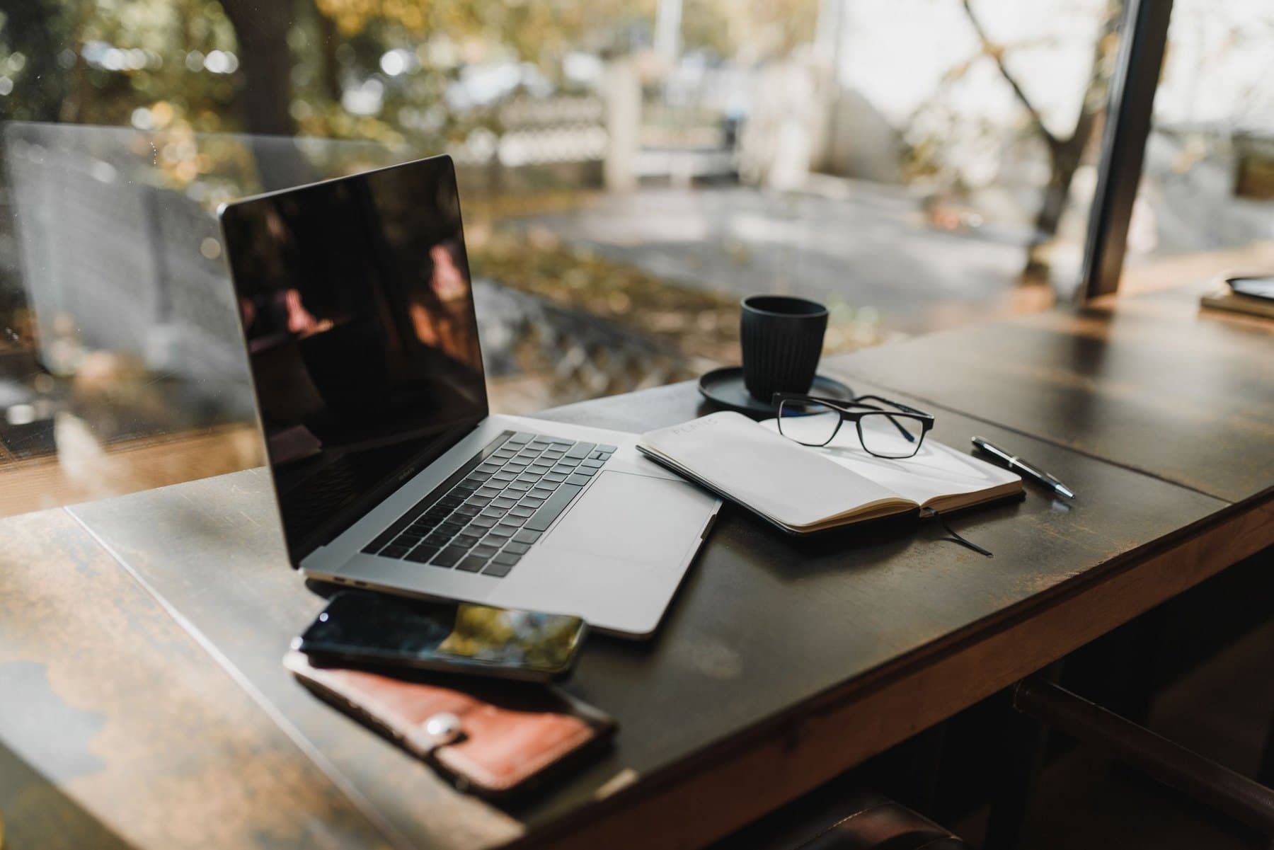 A laptop, an open notebook, and a cup of coffee on a table beside a window
